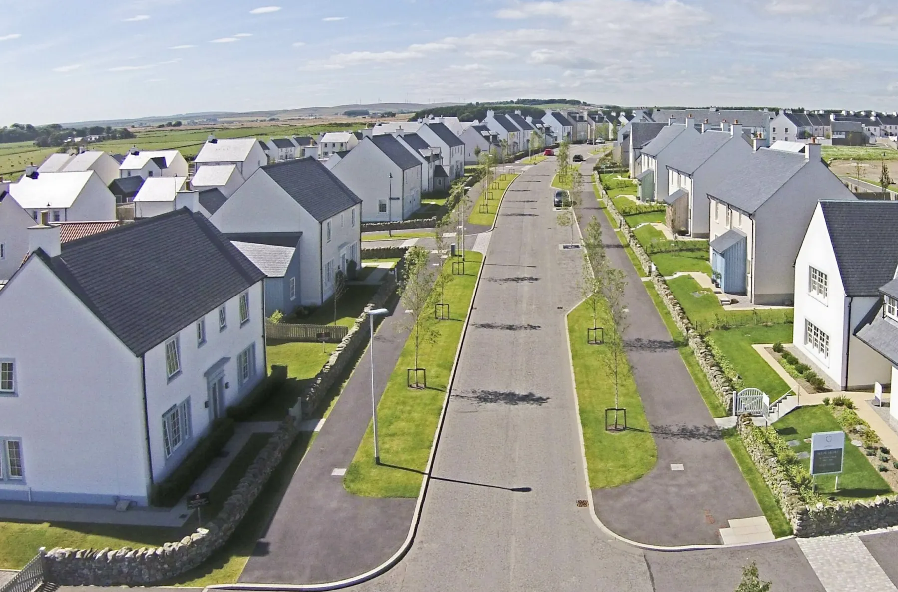 An aerial view of the new town, Chapelton of Elsick. A broad street with grass on either side recedes into distance. Along either side are rows of detached houses of various sizes. Further houses behind are just in shot.
