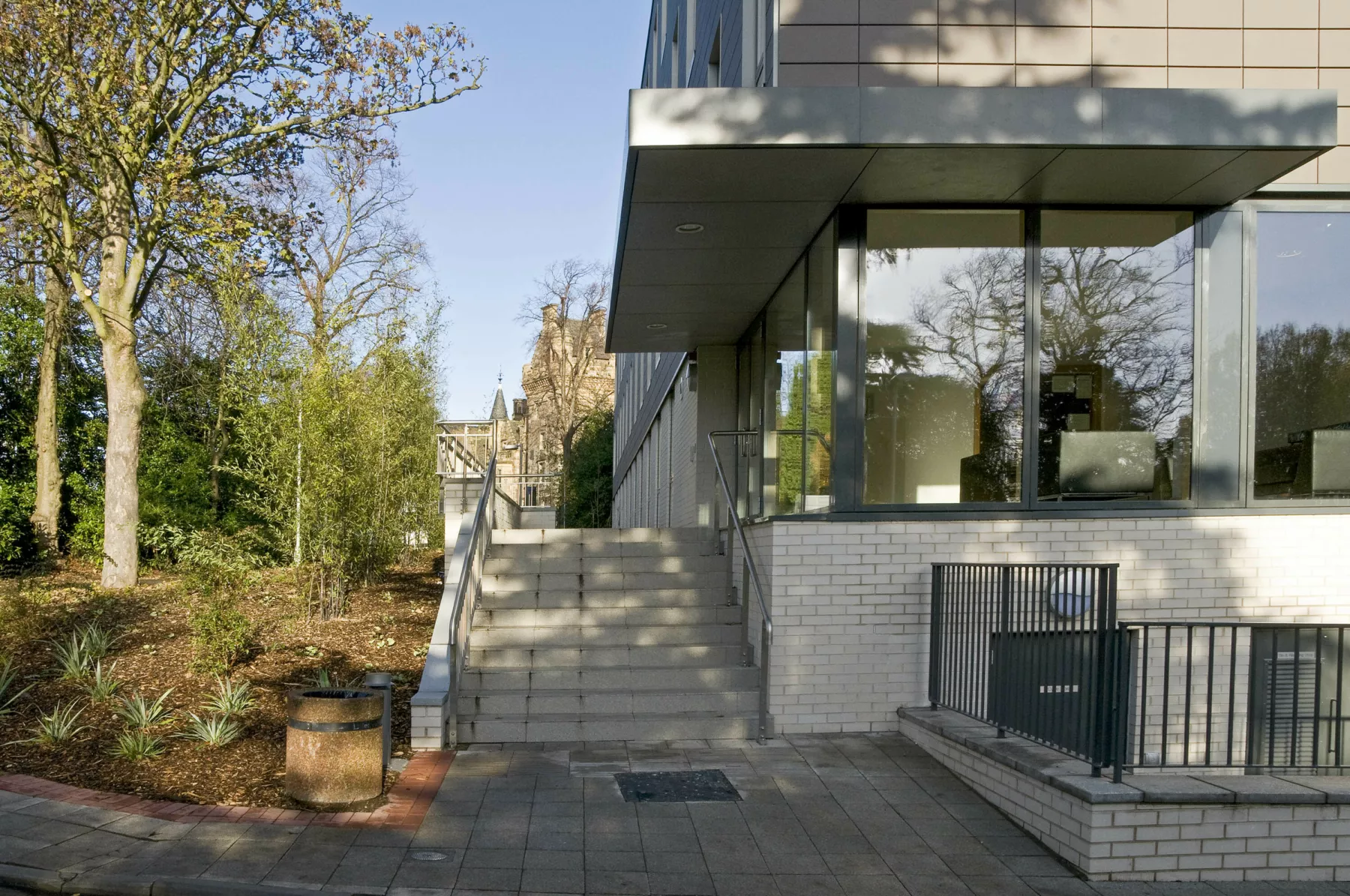 View of the entrance to John Burnett House, student residential block at University of Edinburgh's Pollock Halls of Residence. Floor to ceiling glazed entrance hall within the pale brick clad ground floor. Above, the floors are in bronze-coloured cladding. Landscaped grounds to the left.
