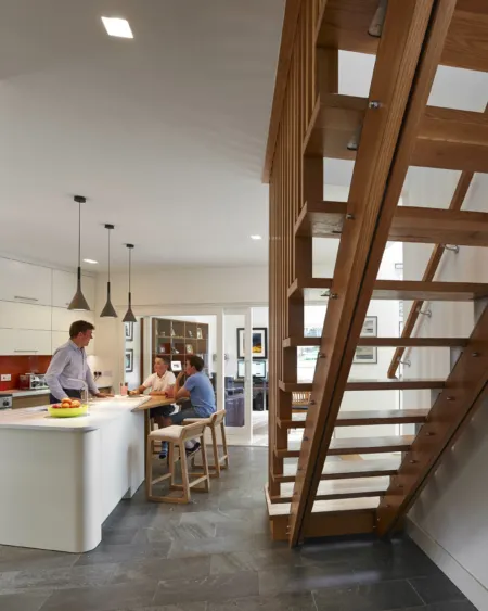 In a new built private house in Edinburgh, An exposed timber staircase with open treads descends into a family kitchen area. Pendant lights are arranged about a white kitchen island with seating. The family is gathered around. The living room can be seen through glass doors.