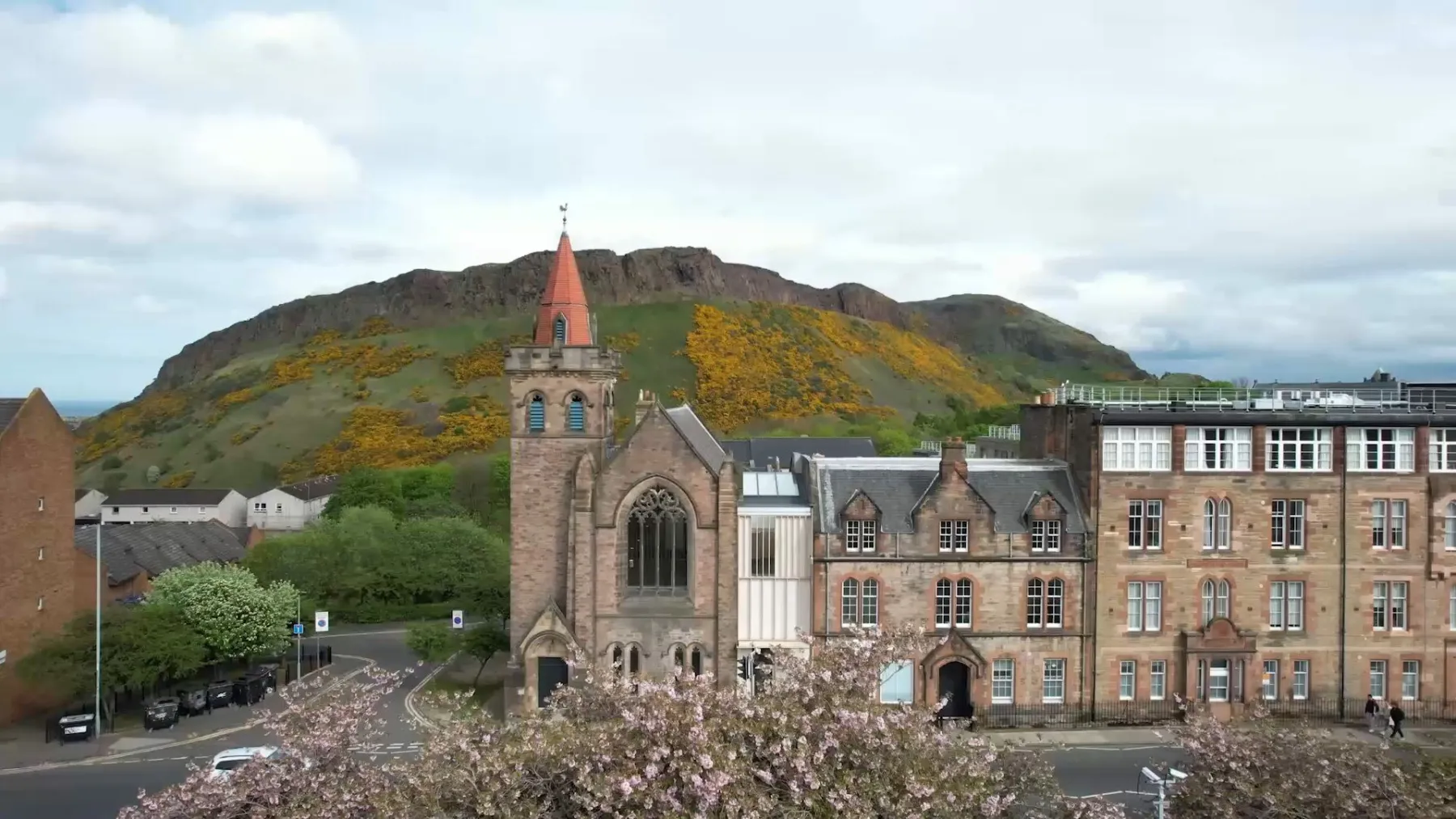 Greyfriars Charteris Centre, Edinburgh. A church building with a contemporary extension inserted between in and further stone-built buildings. Cherry blossom is in the foreground with Arthur's Seat is the background.