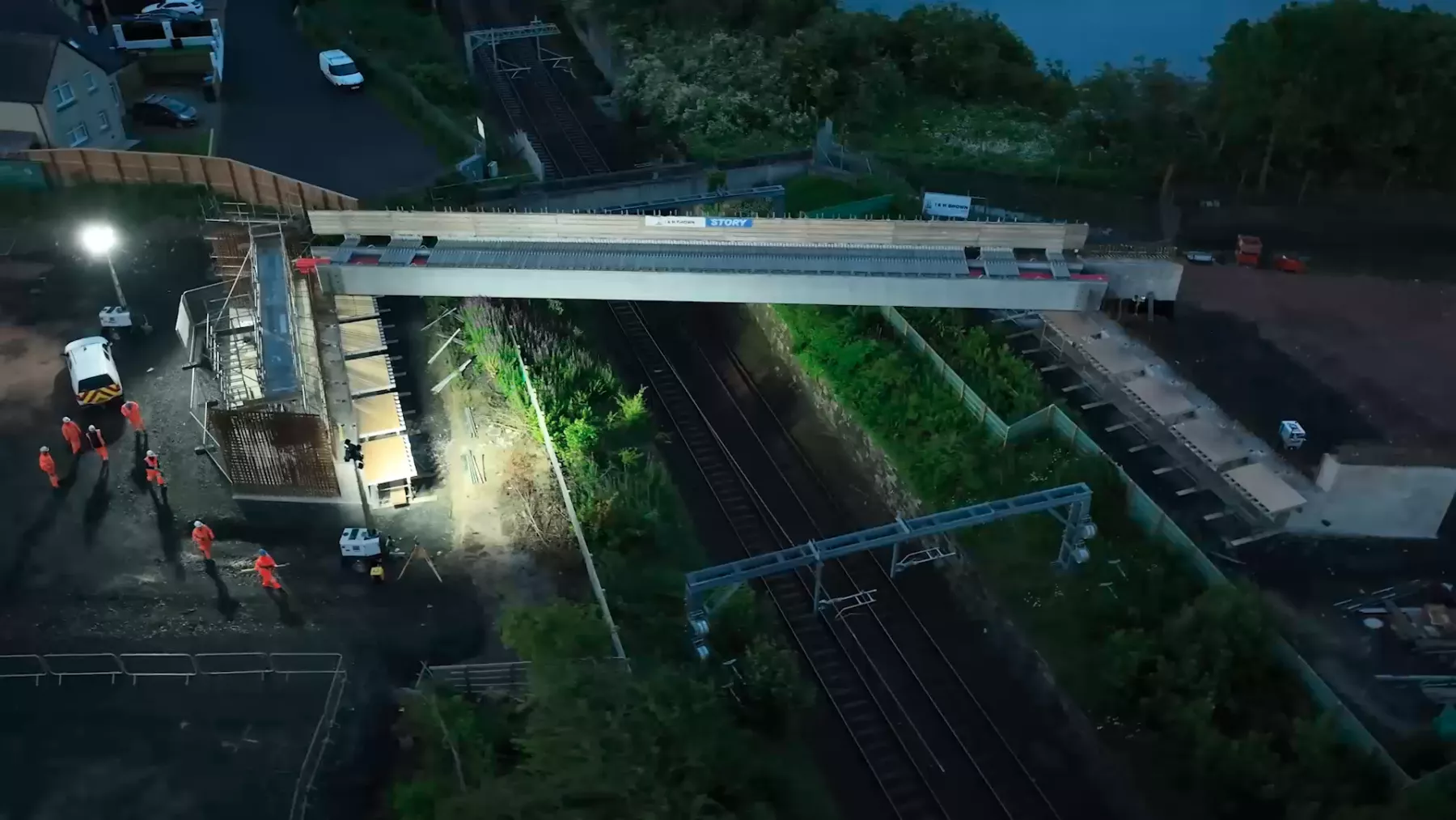 Shot from a drone - workers dressed in orange hi-vis clothing in the very early morning in low light beside the railway line at Winchburgh, West Lothian. A new bridge is being constructed. Floodlights are in use.