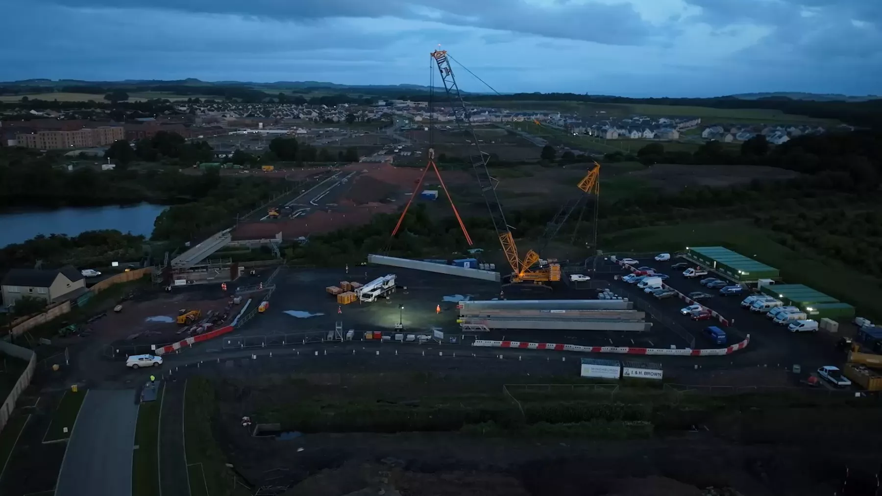 Shot from a drone - workers dressed in orange hi-vis clothing in the very early morning in low light beside the railway line at Winchburgh, West Lothian. A new bridge is being constructed. Floodlights are in use in places as a crane lifts sections of the bridge.